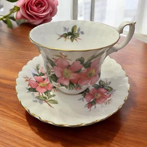 Puede incluir: Un juego de taza y platillo blanco con un patr&oacute;n floral de rosas rosas. La taza y el platillo tienen un borde dorado.
