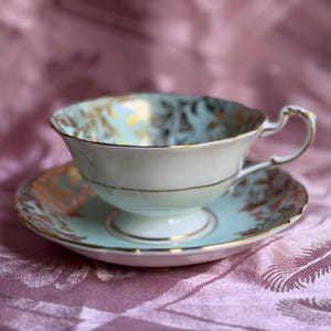 May include: A light blue porcelain teacup and saucer set featuring a delicate gold leaf pattern along the scalloped rims and interior. The teacup has a pedestal base and a curved handle, with gold trim accenting the edges of both pieces.