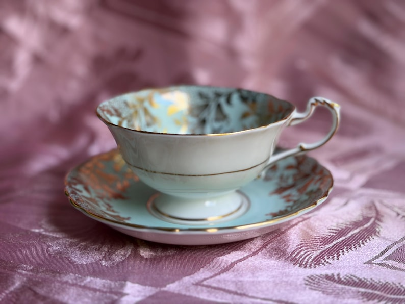 May include: A light blue porcelain teacup and saucer set featuring a delicate gold leaf pattern along the scalloped rims and interior. The teacup has a pedestal base and a curved handle, with gold trim accenting the edges of both pieces.