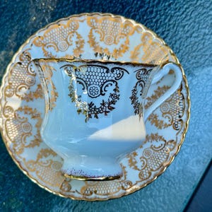 May include: A white porcelain teacup and saucer set featuring an intricate gold filigree pattern. The cup has a scalloped rim and a curved handle, while the saucer displays a matching ornate gold design with a scalloped edge and lace-like details.
