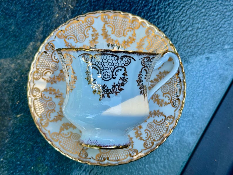May include: A white porcelain teacup and saucer set featuring an intricate gold filigree pattern. The cup has a scalloped rim and a curved handle, while the saucer displays a matching ornate gold design with a scalloped edge and lace-like details.