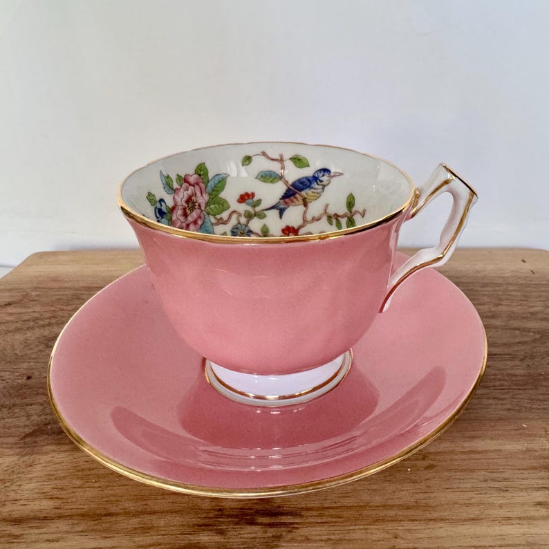 May include: Pink teacup and saucer set with gold trim. The inside of the teacup features a detailed illustration of a blue bird perched on a branch with flowers. The teacup and saucer are made of porcelain.
