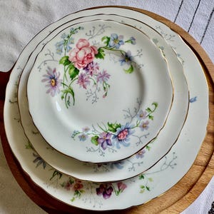 May include: Three white porcelain plates with a floral pattern of pink, purple, and blue flowers, each with a gold rim. The plates are stacked on a wooden serving board.