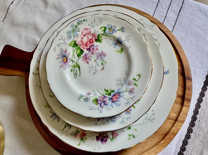 May include: Three white porcelain plates with a floral pattern of pink, purple, and blue flowers, each with a gold rim. The plates are stacked on a wooden serving board.