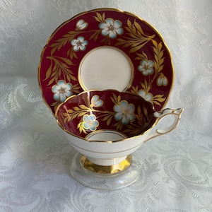 May include: A porcelain teacup and saucer set featuring a deep burgundy background with light blue floral motifs and gold leaf accents. The cup has a scalloped rim, a gold-trimmed handle, and a gold base, while the saucer matches the design.