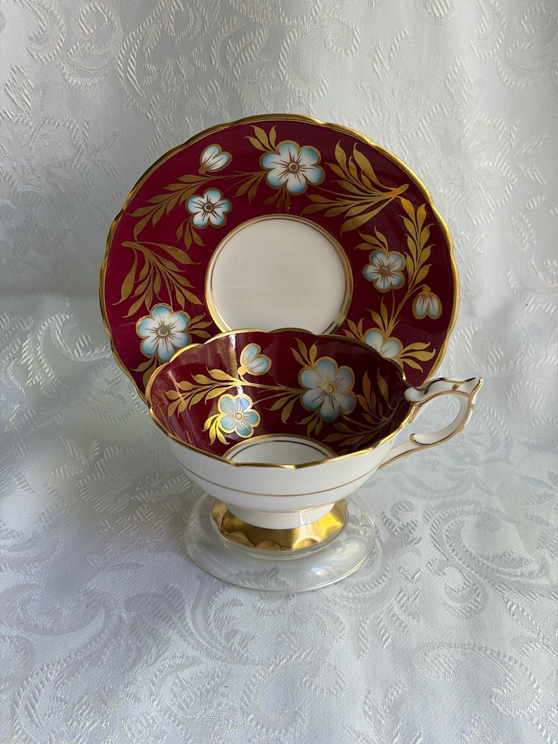 May include: A porcelain teacup and saucer set featuring a deep burgundy background with light blue floral motifs and gold leaf accents. The cup has a scalloped rim, a gold-trimmed handle, and a gold base, while the saucer matches the design.