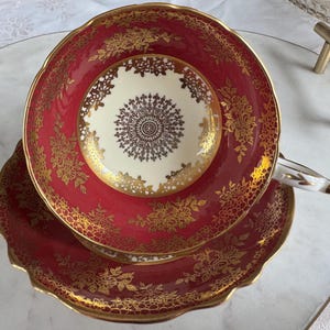 May include: A vintage teacup and saucer set. The cup and saucer are a deep red with intricate gold floral patterns. The center of the cup is cream-colored with a detailed gold design. The set has a scalloped edge and a gold trim.