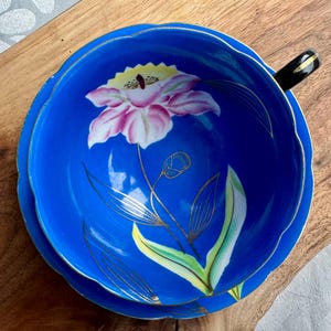 Puede incluir: Una taza de t&eacute; y un platillo azules con un borde dorado y un dise&ntilde;o floral. La flor es rosa con acentos amarillos y est&aacute; pintada sobre un fondo azul.