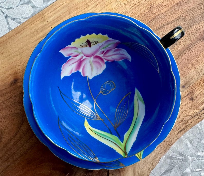 Puede incluir: Una taza de t&eacute; y un platillo azules con un borde dorado y un dise&ntilde;o floral. La flor es rosa con acentos amarillos y est&aacute; pintada sobre un fondo azul.