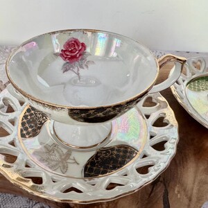 Puede incluir: Juego de taza y platillo blancos con dise&ntilde;o de rosa. La taza tiene asa y borde dorados, y el platillo tiene un dise&ntilde;o calado en forma de coraz&oacute;n. El juego est&aacute; sobre una superficie de madera.