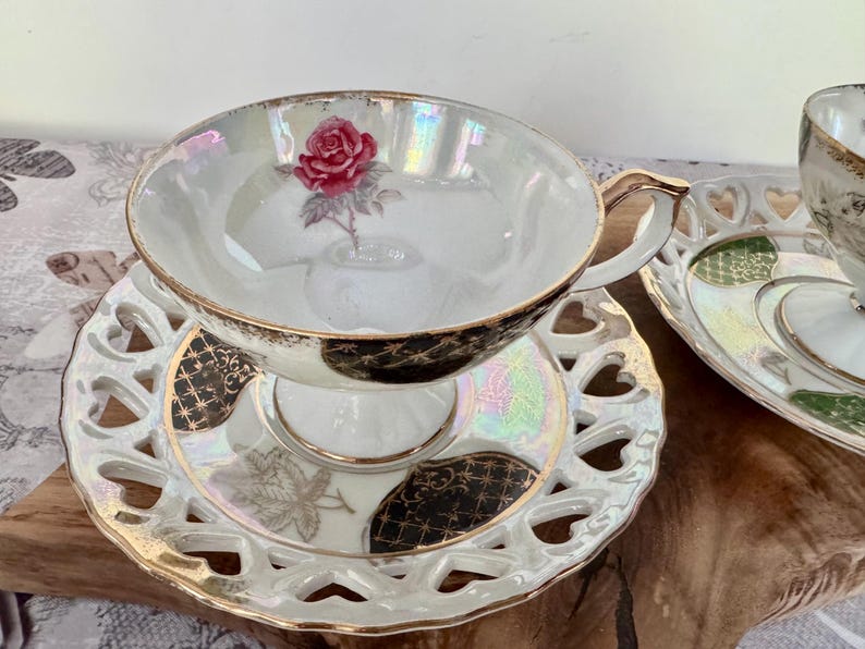 Puede incluir: Juego de taza y platillo blancos con dise&ntilde;o de rosa. La taza tiene asa y borde dorados, y el platillo tiene un dise&ntilde;o calado en forma de coraz&oacute;n. El juego est&aacute; sobre una superficie de madera.