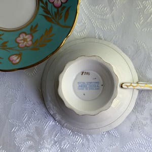 May include: A white bone china teacup and saucer set. The saucer features a teal border with pink flowers and gold leaf accents. The underside of the teacup shows the Royal Stafford Bone China Made in England stamp and the number 8506.