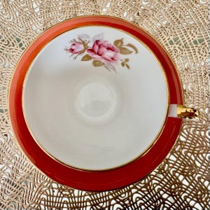 May include: A white teacup with a gold rim and a floral design featuring pink roses and gold leaves. The teacup is set against a red background.