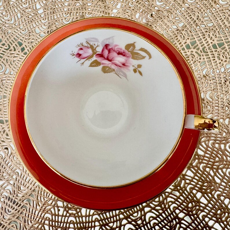 May include: A white teacup with a gold rim and a floral design featuring pink roses and gold leaves. The teacup is set against a red background.