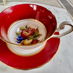 May include: A red teacup and saucer with a gold rim. The teacup has a white interior with a painted design of fruit, including an apple, pear, grapes, and strawberries.
