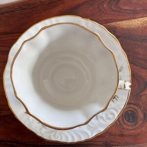 May include: A white teacup and saucer set with a gold rim and floral pattern. The saucer has a scalloped edge.
