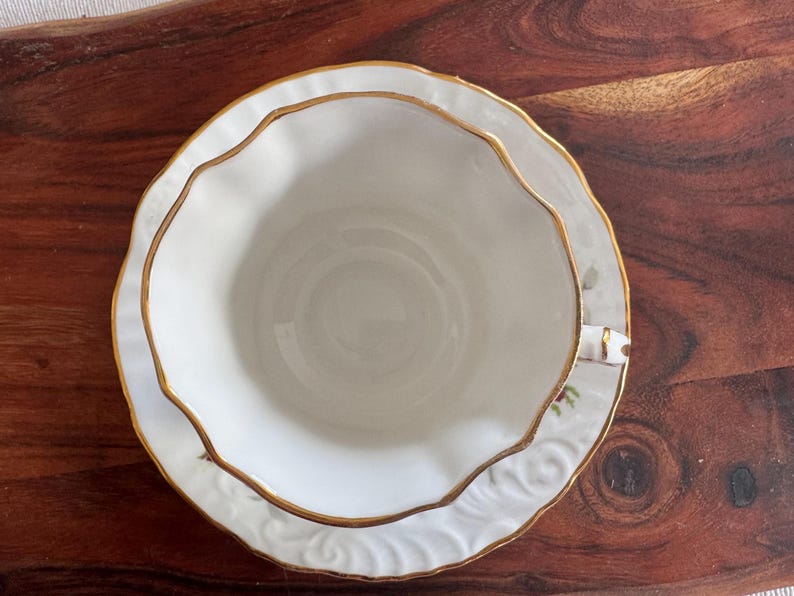 May include: A white teacup and saucer set with a gold rim and floral pattern. The saucer has a scalloped edge.