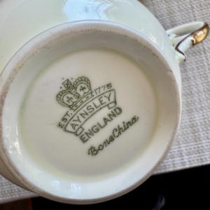 May include: Close-up of the bottom of a white teacup with a green and gold logo that reads "Est. 1775 Aynsley England Bone China".