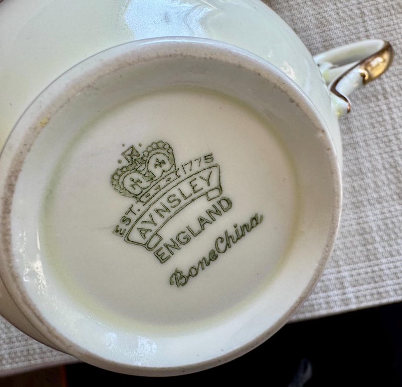 May include: Close-up of the bottom of a white teacup with a green and gold logo that reads "Est. 1775 Aynsley England Bone China".