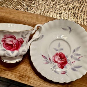 May include: A white teacup and saucer set with a pink rose floral pattern. The cup has a gold rim and the saucer has a scalloped edge.