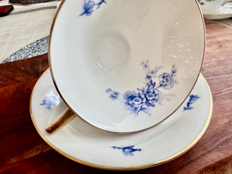 May include: A white teacup and saucer with a blue floral pattern and gold trim. The cup and saucer are both decorated with delicate blue flowers and leaves.