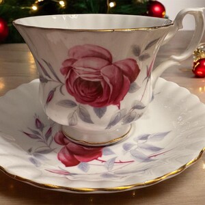 May include: A white teacup and saucer set with a floral design featuring a large pink rose with gray leaves. The set has a gold trim around the edges.