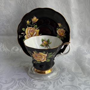 Pode incluir: Um conjunto de ch&aacute;vena e pires preto com ilustra&ccedil;&otilde;es de rosas amarelas e folhas verdes. A ch&aacute;vena tem um interior branco, um rebordo dourado e uma base dourada. O pires apresenta um rebordo recortado com acabamento em ouro e rosas amarelas.
