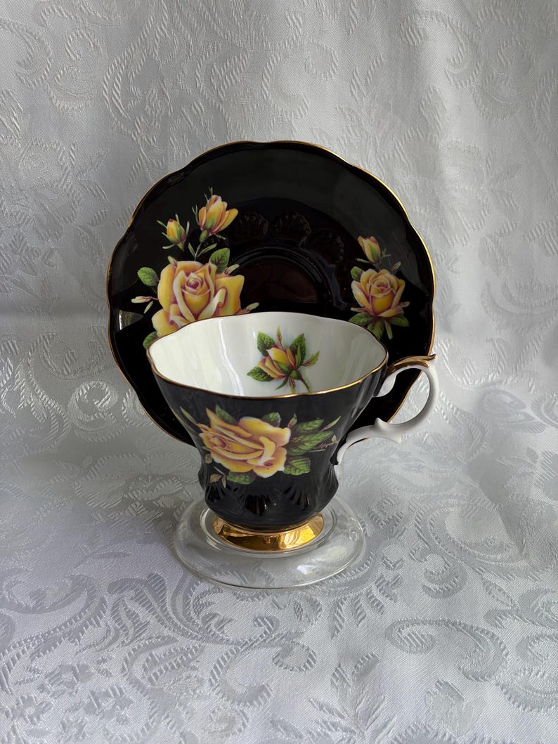 Pode incluir: Um conjunto de ch&aacute;vena e pires preto com ilustra&ccedil;&otilde;es de rosas amarelas e folhas verdes. A ch&aacute;vena tem um interior branco, um rebordo dourado e uma base dourada. O pires apresenta um rebordo recortado com acabamento em ouro e rosas amarelas.