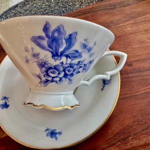 May include: A white porcelain teacup and saucer with a blue floral design. The cup has a scalloped edge and a gold trim. The saucer has a matching floral design.