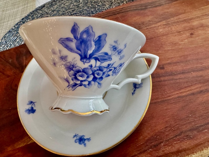 May include: A white porcelain teacup and saucer with a blue floral design. The cup has a scalloped edge and a gold trim. The saucer has a matching floral design.