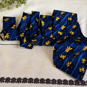 May include: A navy blue necktie with a repeating pattern of yellow floral designs and diagonal blue stripes. The tie is made of a smooth fabric and is displayed on a white surface.