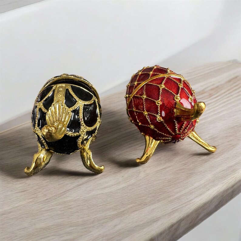May include: Two decorative egg-shaped trinket boxes with gold accents. One box is black with gold detailing and the other is red with gold detailing.