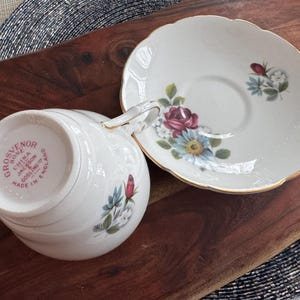 May include: A white teacup and saucer with a floral design. The teacup has the words "Grosvenor Bone China Jackson & Gosling Ltd Made in England" on the bottom.