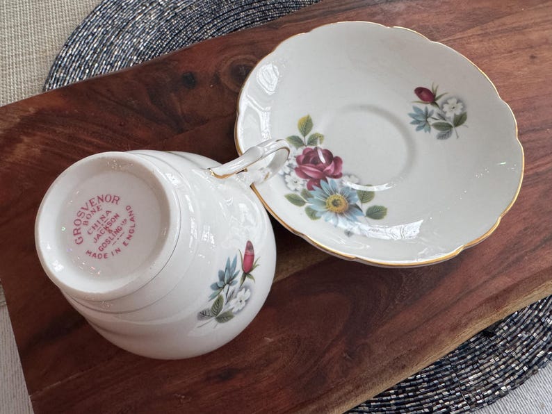 May include: A white teacup and saucer with a floral design. The teacup has the words "Grosvenor Bone China Jackson & Gosling Ltd Made in England" on the bottom.