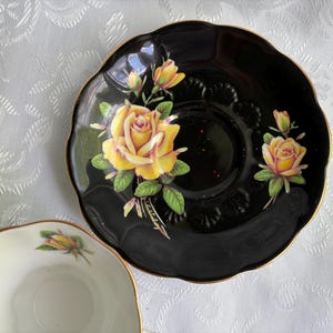 Pode incluir: Um pires preto com borda recortada e acabamento dourado, apresentando um padr&atilde;o floral de rosas amarelas com folhas verdes. Uma x&iacute;cara de ch&aacute; branca parcial com um design de bot&atilde;o de rosa amarelo correspondente &eacute; vis&iacute;vel no canto inferior esquerdo.