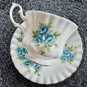 May include: A white teacup and saucer with a blue floral pattern. The cup has a delicate handle and a scalloped rim. The saucer has a gold trim.