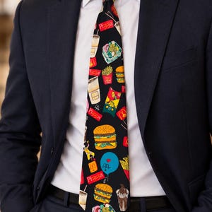 May include: A black necktie featuring a repeating pattern of fast food items including hamburgers, french fries, milkshakes, and blue balloons with hearts. The word Pizza is printed in red text throughout the design on the fabric.