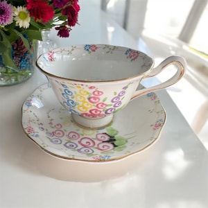 May include: A white teacup and saucer with a floral design. The cup and saucer are decorated with colourful flowers, including pink, purple, yellow, and blue. The cup has a gold rim and a handle.