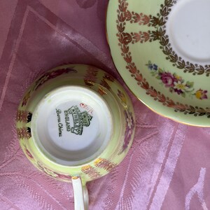 May include: A pale green bone china teacup and saucer set featuring a floral pattern with pink roses and gold leaf accents. The underside of the teacup displays the Aynsley England maker's mark and the text Bone China in black lettering.