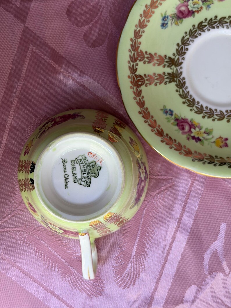 May include: A pale green bone china teacup and saucer set featuring a floral pattern with pink roses and gold leaf accents. The underside of the teacup displays the Aynsley England maker's mark and the text Bone China in black lettering.