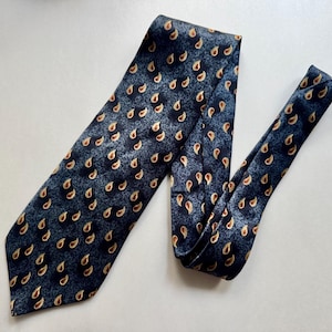 May include: A necktie featuring a dark blue and charcoal gray mottled background with a repeating pattern of small, golden-brown paisley shapes. The tie has a wide, pointed blade at the bottom and a narrower tail section folded over to the right side.
