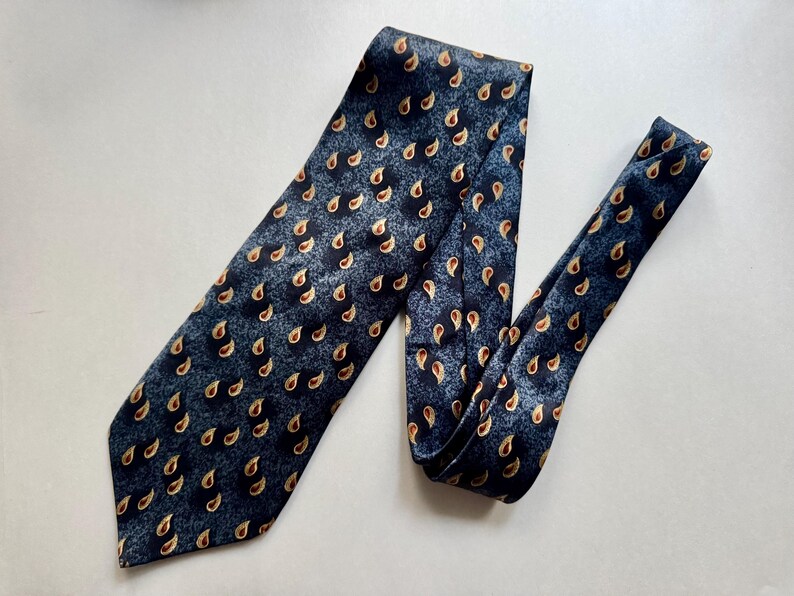 May include: A necktie featuring a dark blue and charcoal gray mottled background with a repeating pattern of small, golden-brown paisley shapes. The tie has a wide, pointed blade at the bottom and a narrower tail section folded over to the right side.