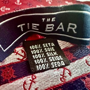 May include: A red silk necktie featuring a repeating pattern of white anchors and ship wheels. The fabric label reads THE TIE BAR, and a smaller black tag indicates 100% SETA, 100% SOIE, 100% SILK, 100% SEIDE, 100% SEDA, confirming the material composition.