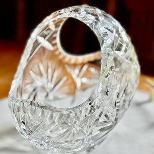 May include: A clear, cut-glass basket with a handle. The basket features intricate starburst and geometric patterns. The handle has a rounded opening. The basket is sitting on a white surface.