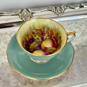 May include: A vintage teacup and saucer set. The cup and saucer are a soft teal color with gold trim. The inside of the cup features a hand-painted design of peaches, grapes, and leaves. The set is placed on a silver tray.
