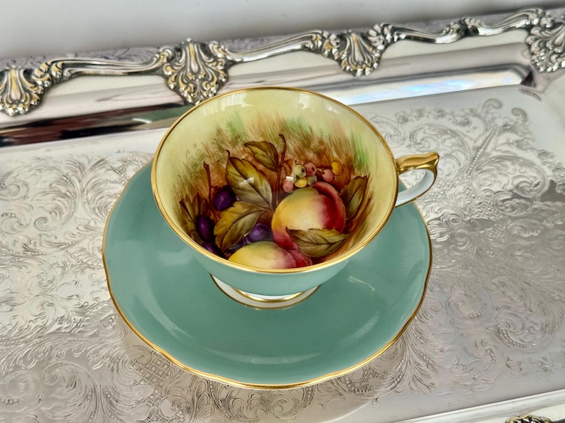 May include: A vintage teacup and saucer set. The cup and saucer are a soft teal color with gold trim. The inside of the cup features a hand-painted design of peaches, grapes, and leaves. The set is placed on a silver tray.