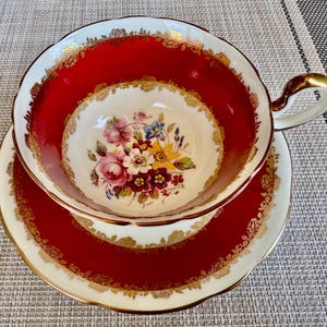 May include: A red and gold teacup and saucer set with a floral design. The cup and saucer have a white background with a red rim and gold trim.