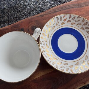 May include: A white teacup and saucer with a blue and gold design. The saucer has a blue center circle with a white inner circle and gold leaf pattern around the edge. The teacup has a gold rim.
