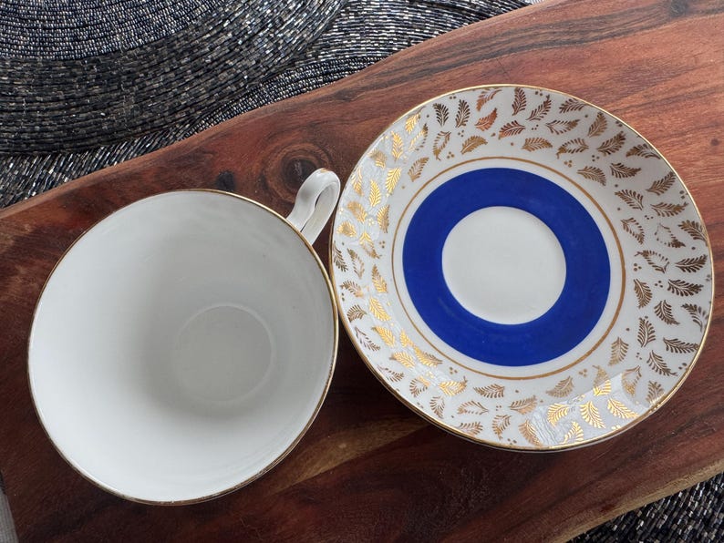 May include: A white teacup and saucer with a blue and gold design. The saucer has a blue center circle with a white inner circle and gold leaf pattern around the edge. The teacup has a gold rim.