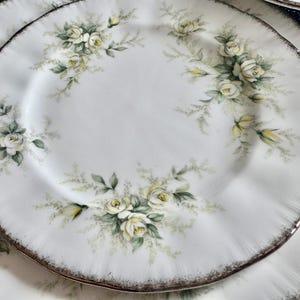 May include: A set of white porcelain plates with a delicate floral design featuring white roses and green leaves. The plates have a gold rim and a scalloped edge.
